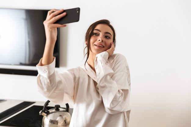 Mujer alegre vistiendo ropa de seda tomando fotos selfie en smartphone negro, mientras está de pie en la cocina por la mañana