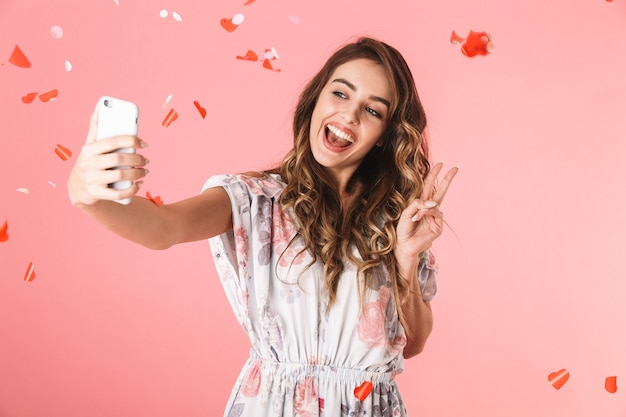 Mujer alegre vistiendo un vestido sonriendo y tomando selfie bajo confeti cayendo, aislado en rosa