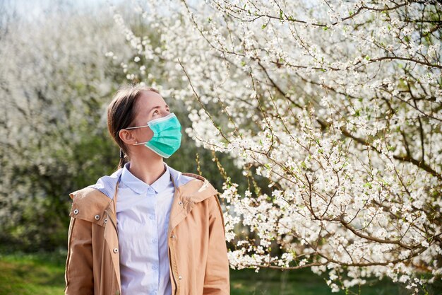 Mujer alérgica que sufre de alergia estacional en primavera posando en un jardín en flor en primavera con una máscara médica mirando un árbol en flor Concepto de alergia de primavera