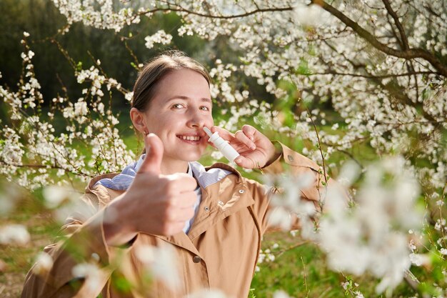 Foto mujer alérgica que sufre de alergia estacional en primavera posando en un jardín en flor en primavera mujer feliz que usa gotas nasales mostrando los pulgares hacia arriba entre los árboles en flor concepto de alergia de primavera