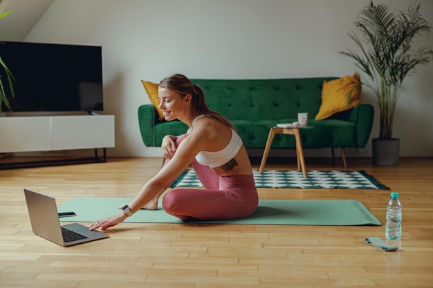 Mujer en una alfombra de yoga con ropa deportiva usando una computadora portátil en un piso de madera dura