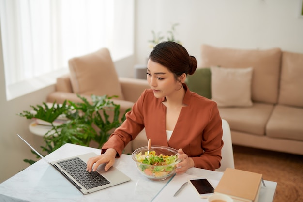 Mujer almorzando en un tazón de vidrio y usando una computadora portátil.