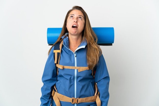 Mujer alpinista de mediana edad con una gran mochila sobre pared aislada mirando hacia arriba y con expresión de sorpresa