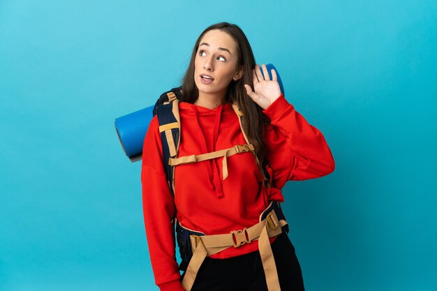 Mujer alpinista con una mochila grande sobre fondo aislado escuchando algo poniendo la mano en la oreja