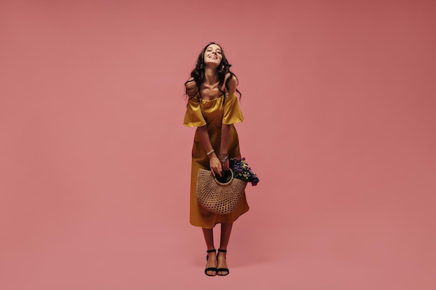 Mujer alta bronceada con cabello rizado en vestido elegante amarillo midi y zapatos negros sonriendo y sosteniendo diadema sobre fondo rosa