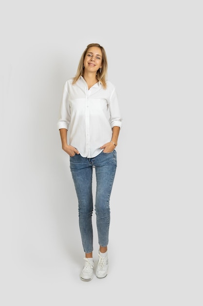 Mujer alta en forma de pie sobre fondo blanco de estudio mirando a la cámara posando con las manos en los bolsillos en blusa blanca jeans azules y zapatillas de deporte Mostrando emociones positivas