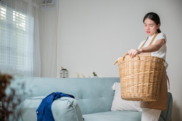 Mujer ama de casa asiática sosteniendo una cesta con un montón de ropa diferente en el sofá de la sala de estar Hermosa ama de casa haciendo tareas domésticas sostiene una cesta de madera de ropa limpia para lavar en casa