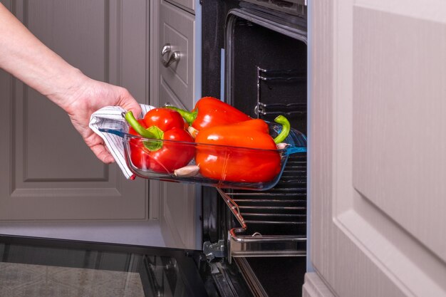 Una mujer ama de casa coloca una bandeja de vidrio ignífugo de pimientos rojos maduros para hornear en un horno eléctrico.