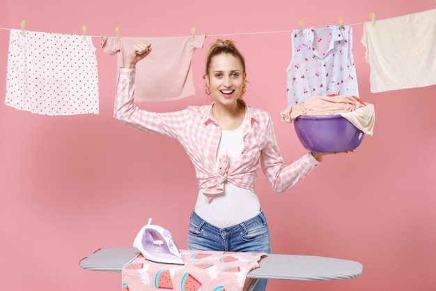 Mujer ama de casa emocionada con camisa a cuadros, ropa seca en la cuerda de planchado a bordo del lavabo con ropa limpia haciendo tareas domésticas aisladas en un fondo rosa. Concepto de limpieza. Mostrando los músculos bíceps.