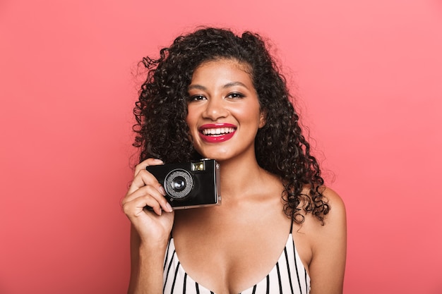 Mujer americana optimista con cabello rizado fotografiando en cámara retro aislado sobre pared rosa