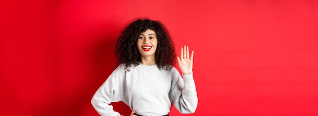 Una mujer amistosa con el pelo rizado te saluda renunciando a la mano y diciendo hola, se presenta de pie