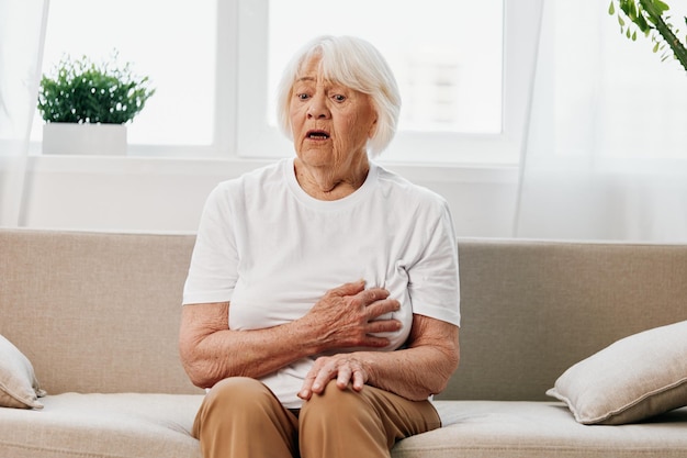 Mujer anciana dolor severo en el corazón sentada en el sofá problemas de salud en la vejez mala calidad de vida abuela con cabello gris sosteniendo su pecho ataque cardíaco y accidente cerebrovascular problema de presión