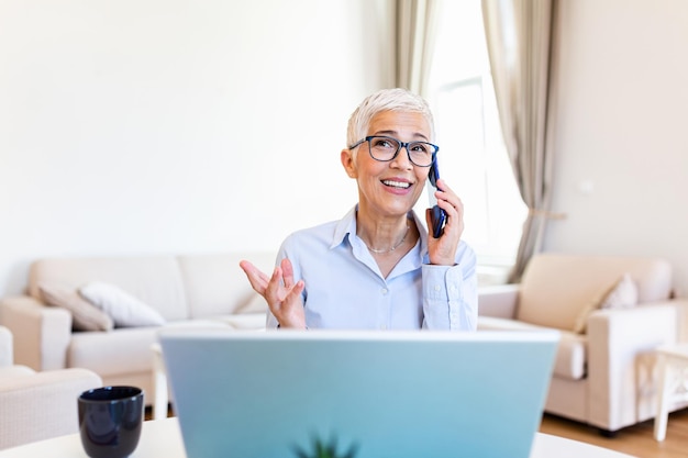 Mujer anciana enfocada con cabello blanco en casa haciendo una llamada de negocios astuta usando una computadora portátil Emprendedora sénior elegante con gafas trabajando en casa Mujer administrando facturas domésticas y finanzas del hogar