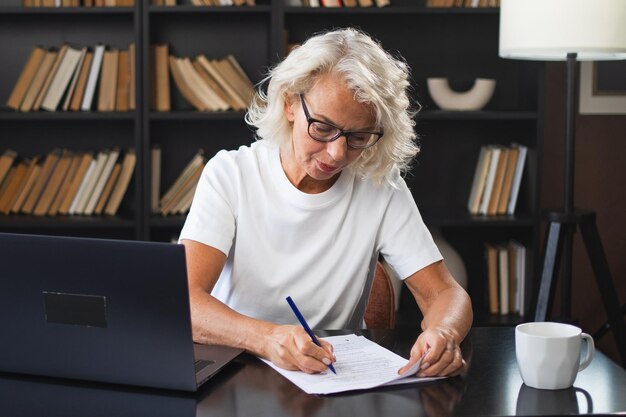 Foto mujer anciana de mediana edad que usa una computadora portátil escribiendo notas en el lugar de trabajo centrada en ancianos maduros