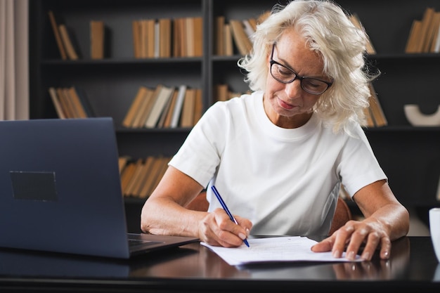 Mujer anciana de mediana edad que usa una computadora portátil escribiendo notas en el lugar de trabajo centrada en ancianos maduros