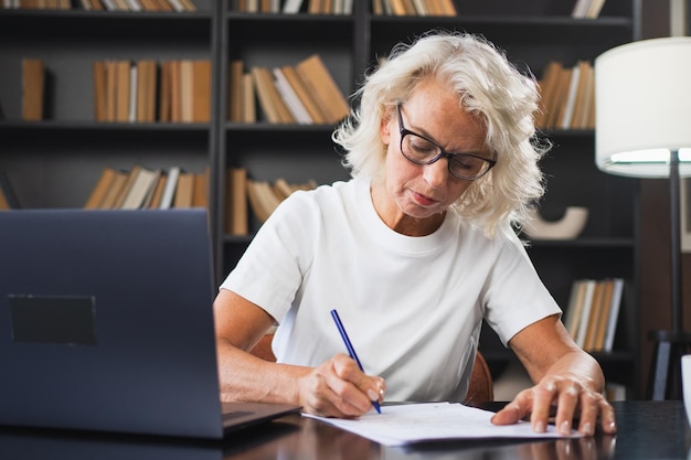Mujer anciana de mediana edad que usa una computadora portátil escribiendo notas en el lugar de trabajo centrada en ancianos maduros