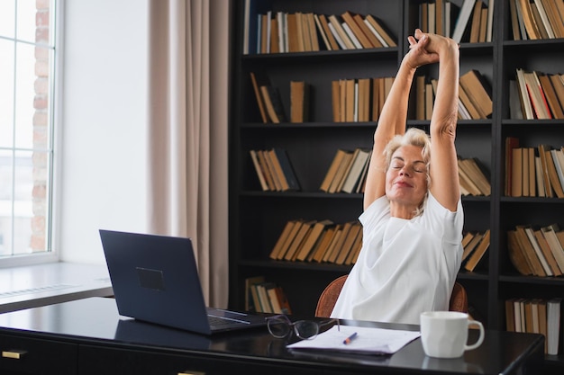 Mujer anciana de mediana edad relajándose estirando los brazos y el cuerpo tomando un descanso del trabajo en la computadora portátil en casa