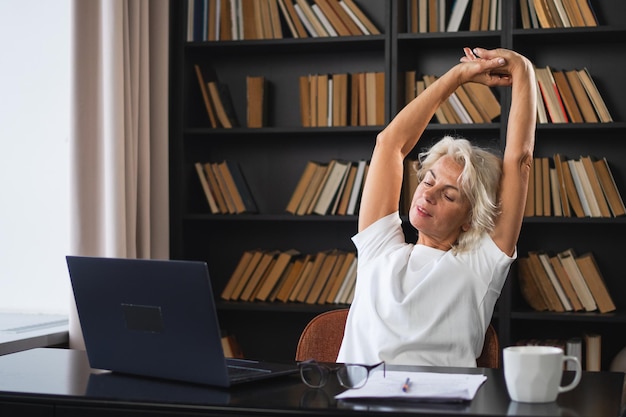 Mujer anciana de mediana edad relajándose estirando las manos y el cuerpo tomando un descanso del trabajo en la computadora portátil en casa