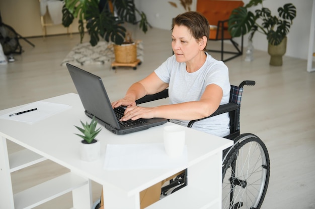 Mujer anciana positiva en silla de ruedas que trabaja en una computadora portátil independiente de TI en línea