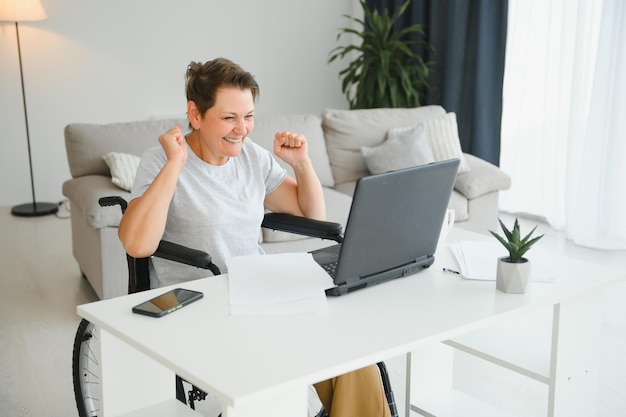 Mujer anciana positiva en silla de ruedas que trabaja en una computadora portátil independiente de TI en línea