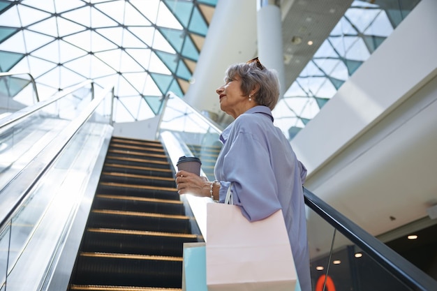 Mujer anciana con ropa casual con café para llevar en la escalera mecánica