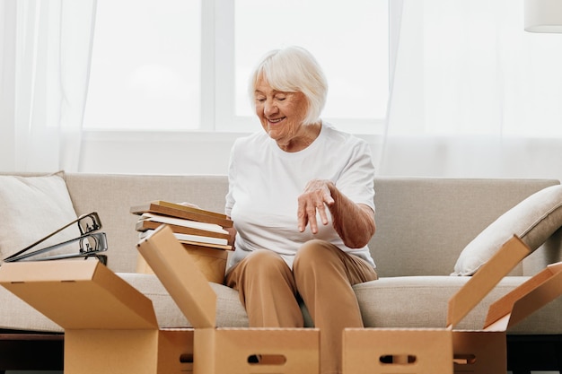 Mujer anciana se sienta en un sofá en casa con cajas recolectando cosas con recuerdos álbumes con fotos y marcos de fotos moviéndose a un nuevo lugar limpiando cosas y una sonrisa feliz estilo de vida jubilación