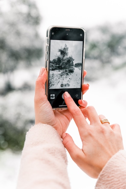 Mujer anónima de cultivo tomando fotos de bosques de invierno cubiertos de nieve mientras usa el teléfono inteligente