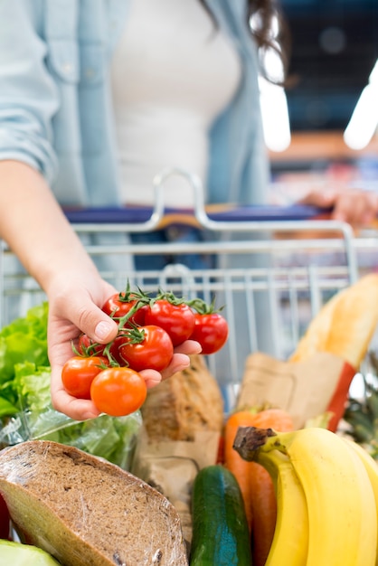 Foto mujer anónima que sostiene los tomates que se colocan con el carro de la compra en el supermercado