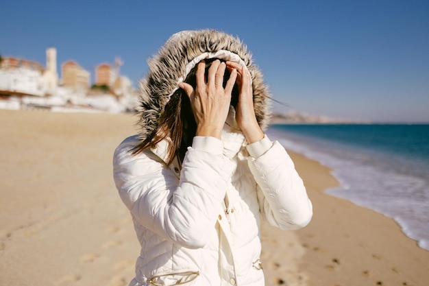 Mujer con anorak blanco escondiendo su rostro con las manos en un soleado día de invierno en la playa