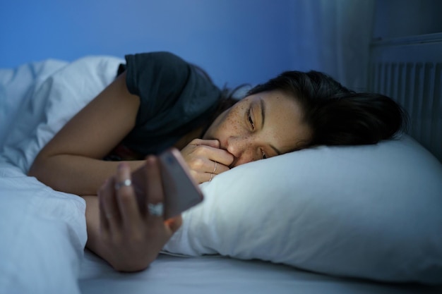 Mujer ansiosa en la cama usando teléfono inteligente por la noche no puede dejar de desplazarse por las redes sociales antes de acostarse