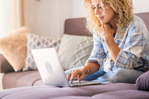 Mujer con anteojos y cabello rizado viendo contenido multimedia usando una computadora portátil en un cómodo sofá en casa. Mujer que trabaja en la computadora portátil en casa. Hermosa mujer con laptop sentada en el sofá en casa