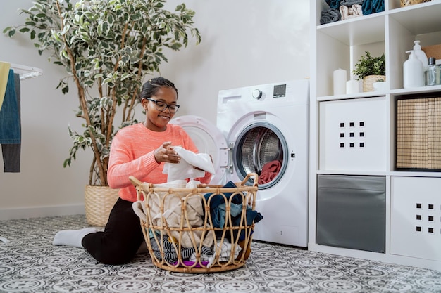 Una mujer con anteojos realiza tareas domésticas en el cuarto de lavado del baño se arrodilla con una canasta de mimbre llena de ropa en la lavadora carga cosas coloridas en el tambor que gira y enjuaga
