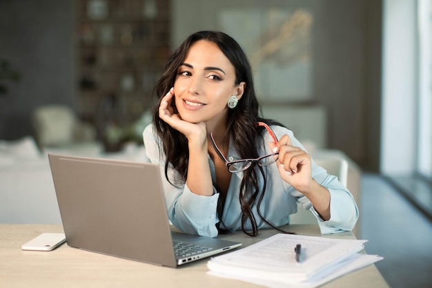 Mujer con anteojos usando una computadora portátil en la oficina en casa trabajando con notas de papel