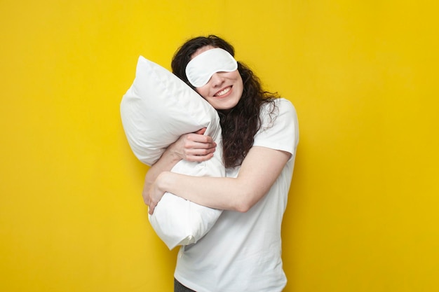 Mujer con antifaz para dormir abraza una almohada suave blanca sobre fondo amarillo y duerme