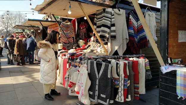 Una mujer de apariencia asiática mira los productos en una tienda de la calle En la ventana del puesto hay hermosos suéteres de punto chalecos abrigos Feria de recuerdos