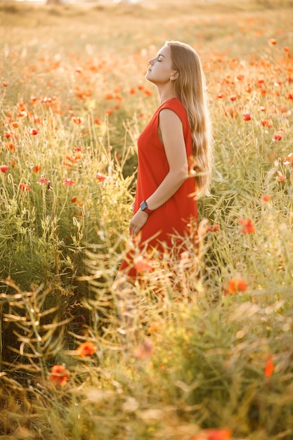 Una mujer de apariencia europea con largo cabello rubio y un vestido rojo de verano, se encuentra en un campo de amapolas en flor