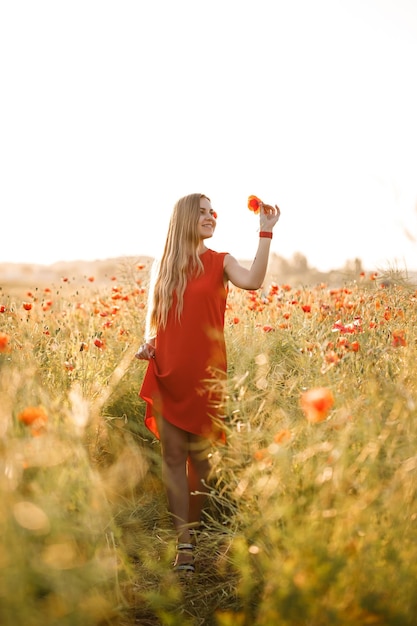 Una mujer de apariencia europea con largo cabello rubio y un vestido rojo de verano, se encuentra en un campo de amapolas en flor