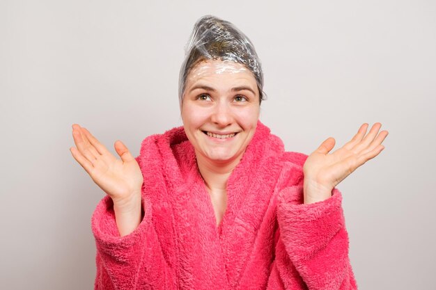Una mujer se aplica una máscara en el cabello y envuelve una película alrededor de su cabeza sonriendo con una bata rosa El concepto de cuidado del cabello