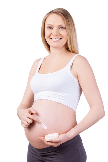 Mujer aplicando crema hidratante. Feliz mujer embarazada aplicando crema hidratante en su vientre de embarazada y sonriendo mientras está de pie aislado en blanco