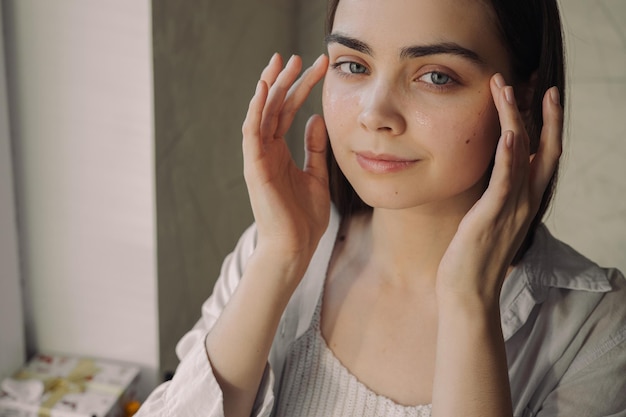 mujer aplicando crema para los ojos y mirando la cámara durante el tratamiento de belleza diario para el cuidado de la piel