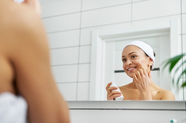 La mujer aplicando productos para el cuidado de la piel facial mientras mira su reflejo en el espejo del baño