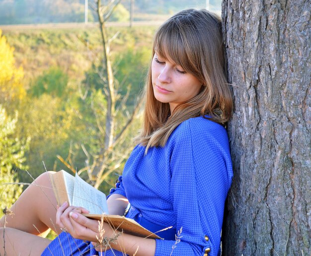 Una mujer apoyada en un árbol leyendo un libro.