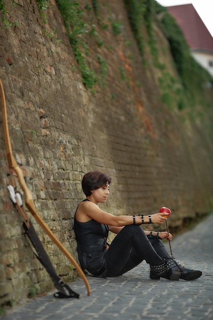 Foto mujer apoyada en la pared de ladrillo cerca de arco y flecha.