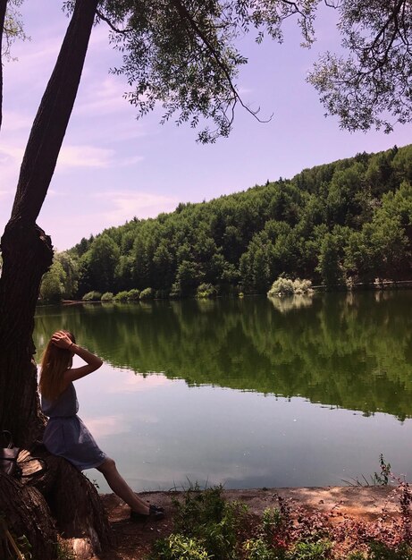 Mujer apoyada en el tronco de un árbol junto al lago contra el cielo