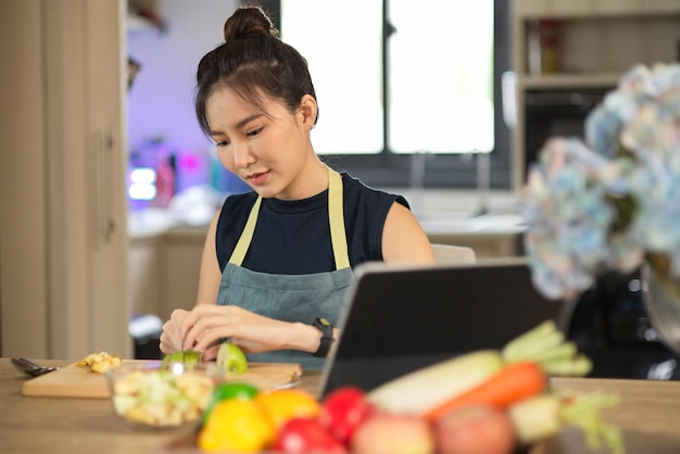 La mujer aprende a cocinar en línea a través de una tableta mientras corta manzanas en una tabla de cortar en la cocina