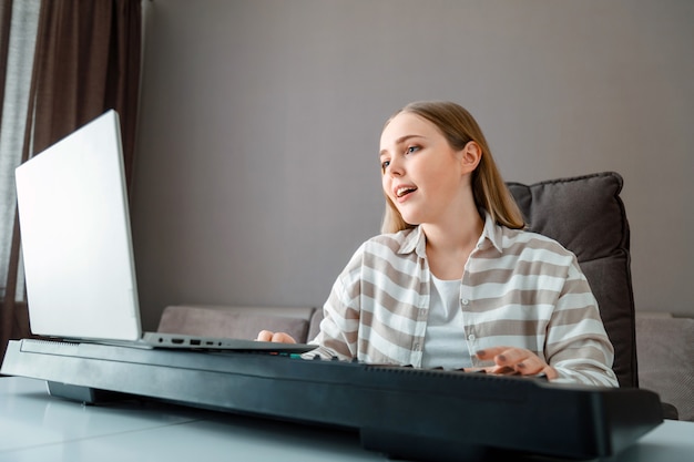 La mujer aprende música cantando voces tocando el piano en línea usando la computadora portátil en el interior de la casa. Chica adolescente canta una canción y toca el sintetizador de piano durante la videollamada, lección en línea con el maestro.