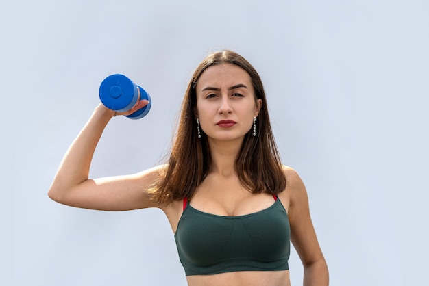 Mujer apta joven en paño verde del deporte sosteniendo pesas aisladas en la pared gris. estilo de vida saludable