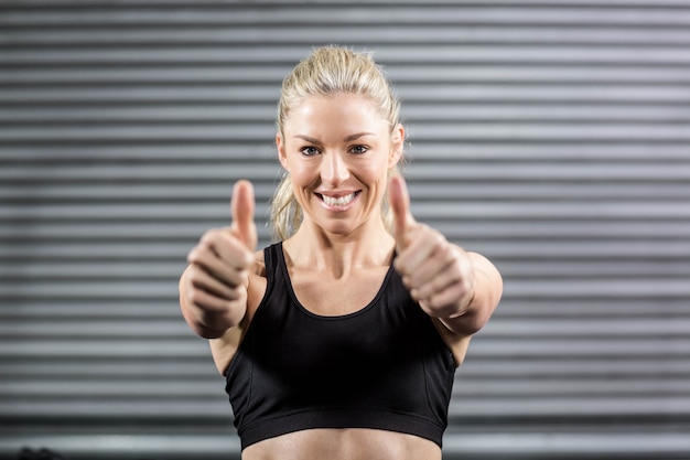 Mujer apta que muestra los pulgares para arriba en el gimnasio de crossfit