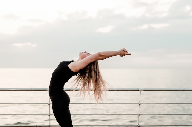 Mujer de la aptitud que hace yoga y que ejercita asana cerca del mar en salida del sol