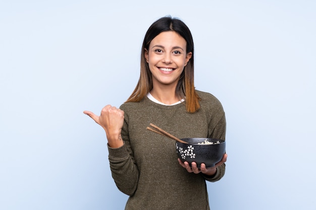 Mujer apuntando hacia un lado para presentar un producto mientras sostiene un tazón de fideos con palillos